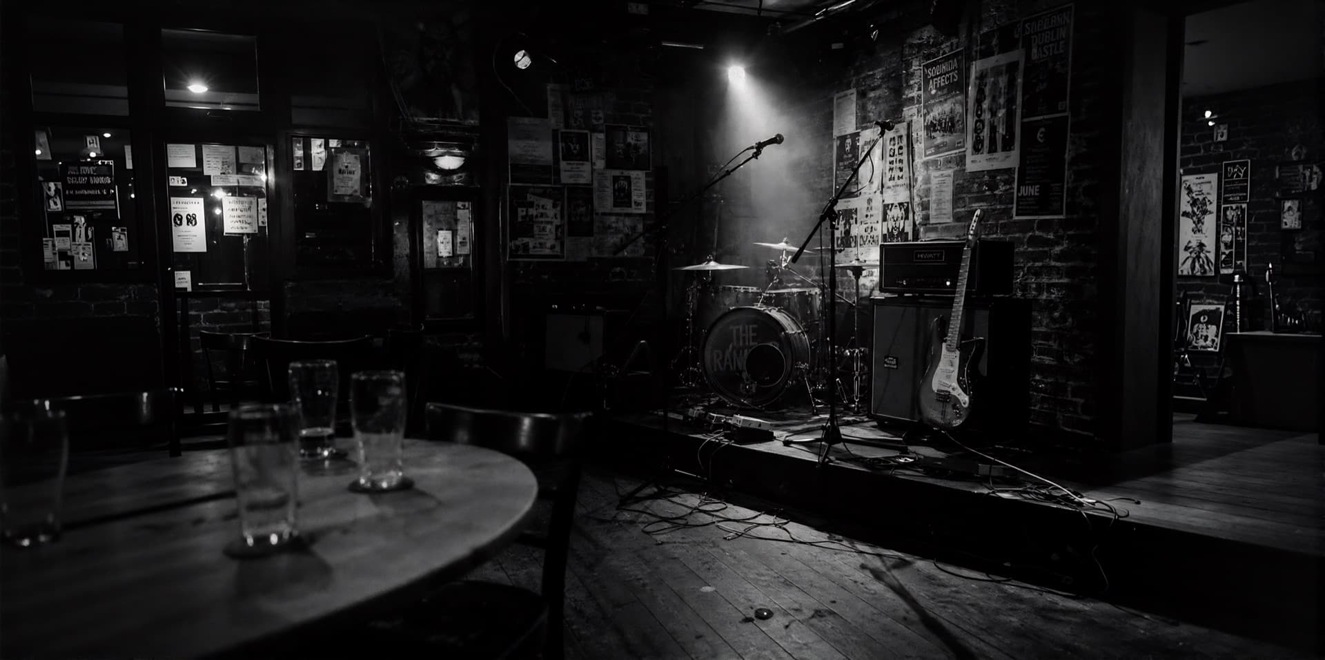 An empty pub band stage at night lit by a single spotlight, drum kit and guitar waiting